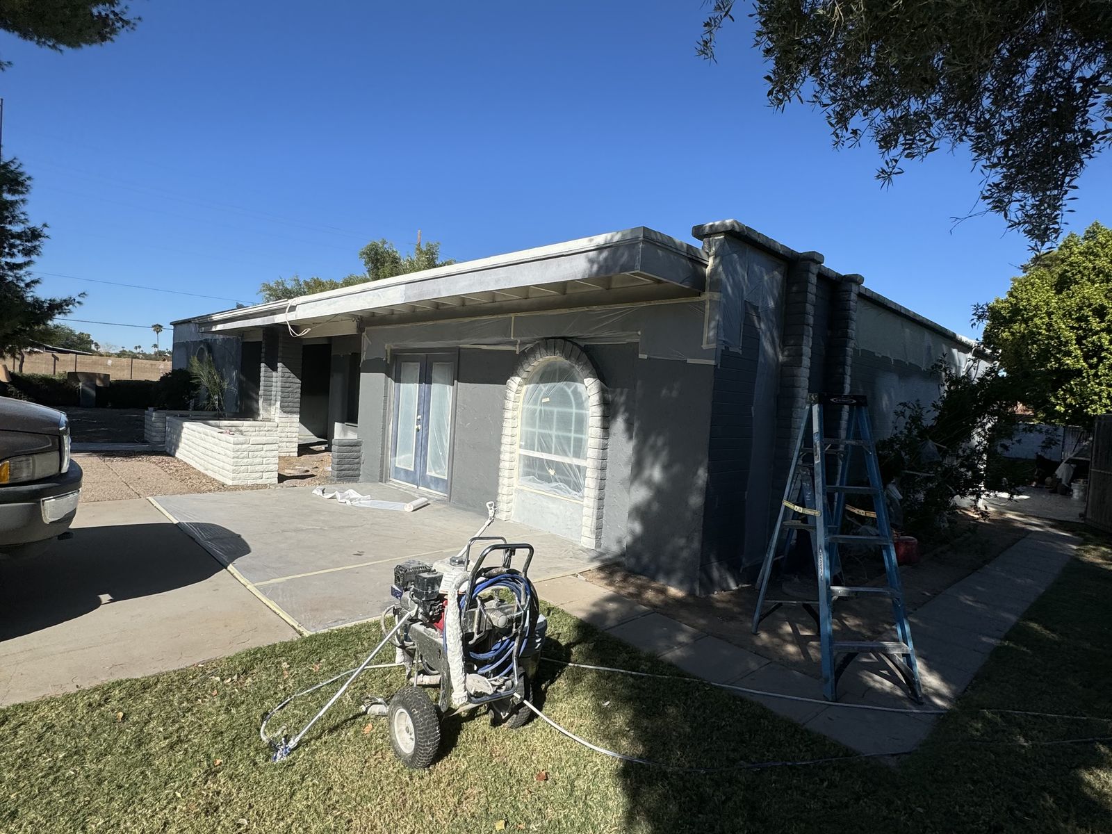 Phoenix block home part-way through an exterior repaint, in grey primer with a sprayer on the lawn